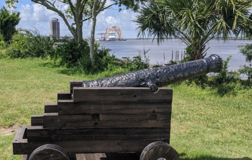 A wooden cannon on wheels, set against a backdrop of water and trees, with buildings in the distance.