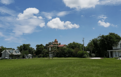 A green field with a historic house and trees under a blue sky with fluffy clouds.