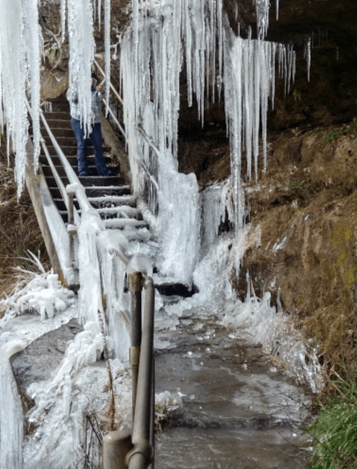 Icicles hang from a rocky overhang near a frozen staircase, with a person standing at the top.