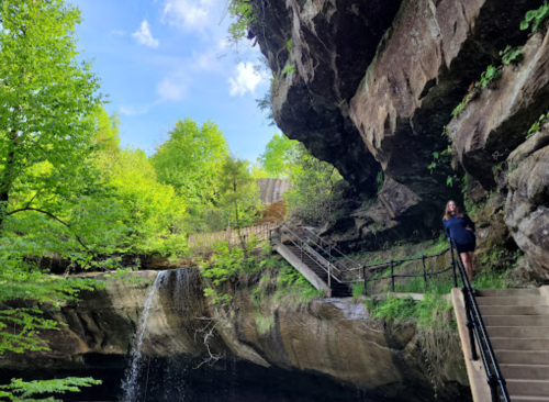 A person stands on a staircase beside a waterfall, surrounded by lush greenery and rocky cliffs under a blue sky.
