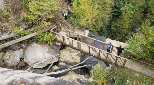 A wooden bridge crosses a rocky area with flowing water, surrounded by trees and autumn foliage.