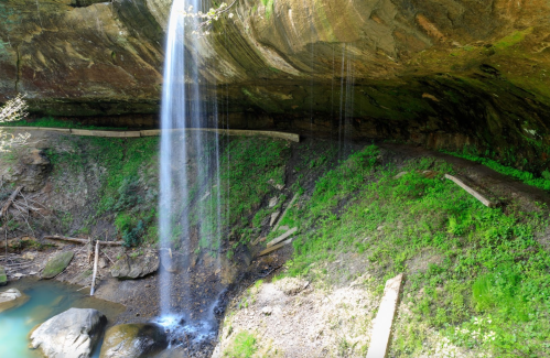 A serene waterfall cascades from a rocky overhang into a lush, green landscape with a winding path nearby.