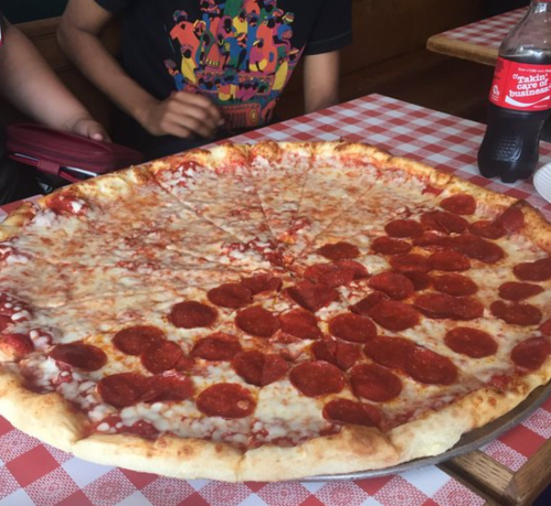 A large pizza topped with pepperoni, served on a checkered tablecloth, with a person partially visible in the background.