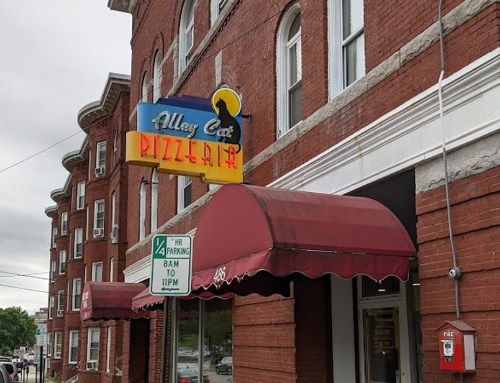 Sign for Alley Cat Pizzeria on a brick building, with red awnings and a parking sign nearby.
