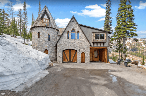 A stone house with a turret, surrounded by snow and pine trees, set against a mountainous landscape.