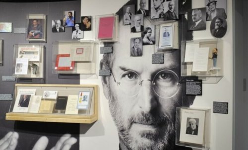 A wall display featuring photos, documents, and artifacts related to notable figures, including a large image of Steve Jobs.