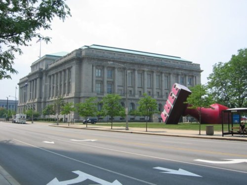 A large red sculpture resembling a giant lipstick stands in front of a historic building on a city street.