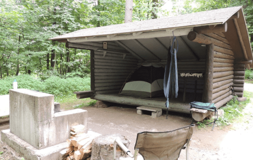 A rustic camping shelter with a tent, firewood, and a picnic area surrounded by lush green trees.