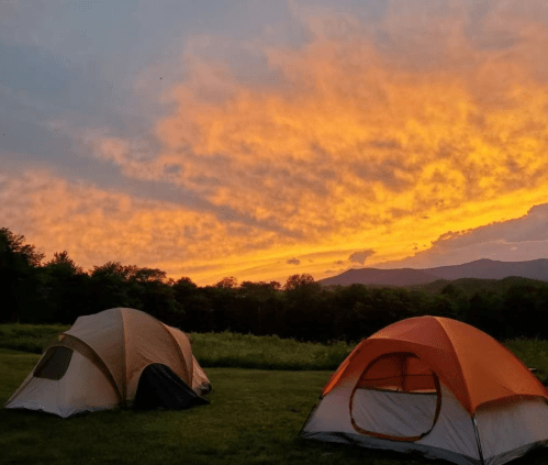 Two tents sit on green grass under a vibrant sunset with colorful clouds and distant mountains in the background.