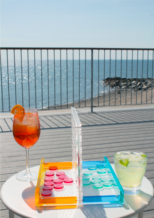 A colorful backgammon set and two cocktails on a table by the seaside, with sparkling water in the background.