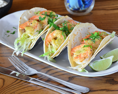 Three shrimp tacos topped with lettuce and cilantro, served on a white plate with lime wedges.