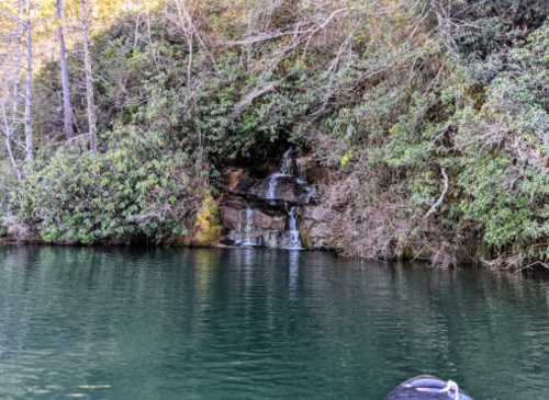 A serene water scene featuring a small waterfall cascading over rocks, surrounded by lush greenery and calm water.