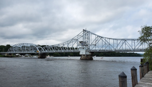 A steel bridge spans a river, with boats visible below and cloudy skies above.