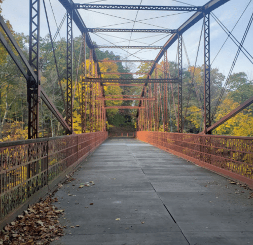 A rusty metal bridge spans a tree-lined path, surrounded by autumn foliage in warm colors.