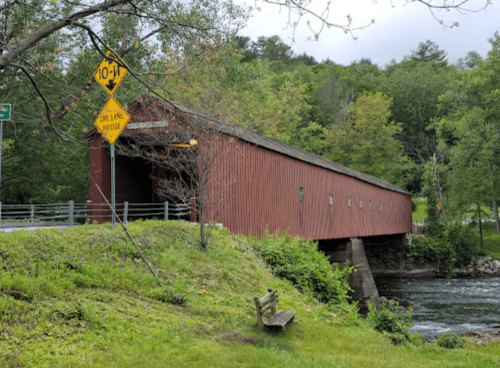 A red covered bridge spans a river, surrounded by green trees and a grassy area, with a warning sign nearby.