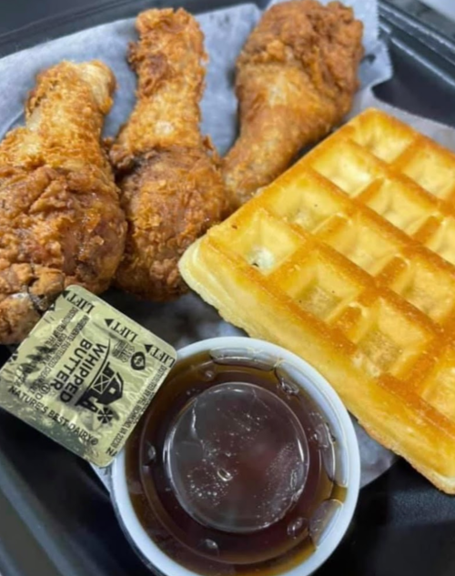 A plate with three fried chicken drumsticks, a waffle, a cup of syrup, and a packet of butter.