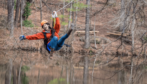 A person in a helmet and harness zip-lining over a calm lake, surrounded by trees in a wooded area.