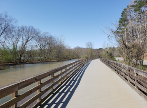 A wooden bridge stretches over a calm river, surrounded by trees and a clear blue sky.