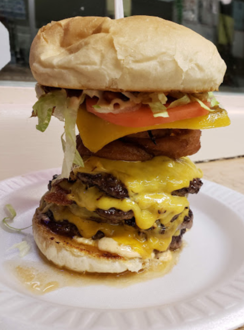 A towering cheeseburger with multiple beef patties, melted cheese, lettuce, tomato, and onion rings on a bun.