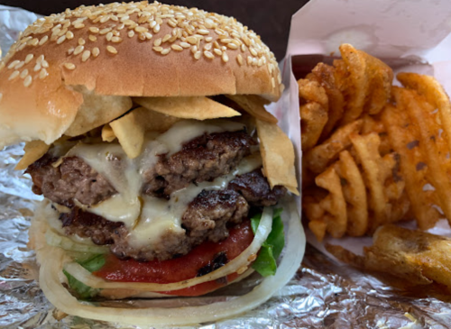 A sesame seed bun burger stacked with two patties, cheese, lettuce, tomato, and tortilla chips, served with waffle fries.
