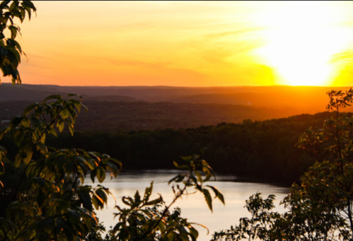 A serene sunset over a calm lake, framed by lush greenery and distant hills.