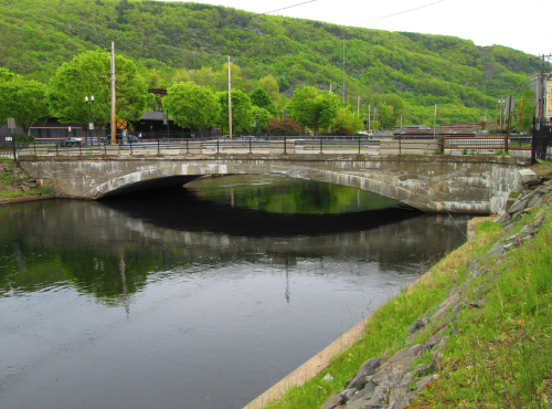 A concrete bridge spans a calm river, surrounded by lush green hills and trees under a cloudy sky.