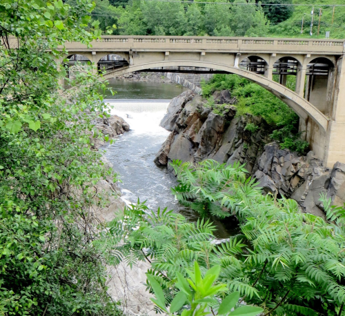 A stone bridge arches over a river, surrounded by lush greenery and rocky cliffs. Water flows beneath the bridge.