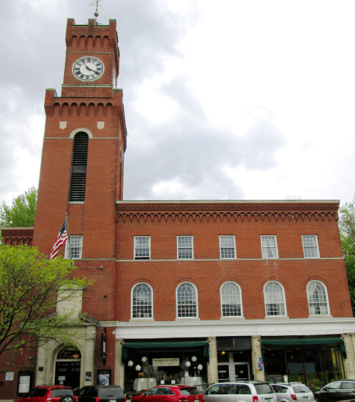Historic brick clock tower building with large windows and an American flag, set against a cloudy sky.