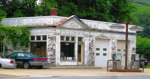 Abandoned gas station with peeling paint, overgrown vines, and old gas pumps, set in a rural area.