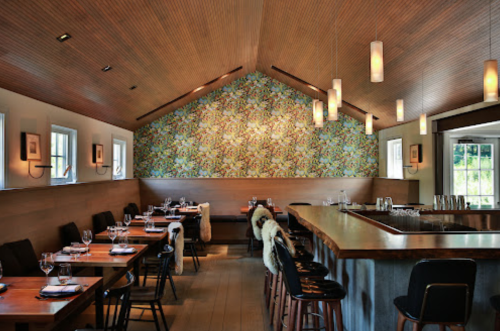 A modern restaurant interior with wooden tables, stylish chairs, and a vibrant floral wallpaper. Soft lighting enhances the ambiance.