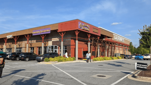 Exterior view of a LA Fitness gym with a parking lot, surrounded by greenery and a clear blue sky.