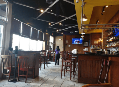 Interior of a restaurant featuring wooden bar stools, a bar area, and a TV, with a person walking in the background.