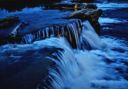 A serene waterfall cascades over rocks, surrounded by dark blue water and a tranquil atmosphere.