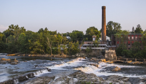 A serene river scene with a stone chimney, lush trees, and gentle rapids under a clear sky.