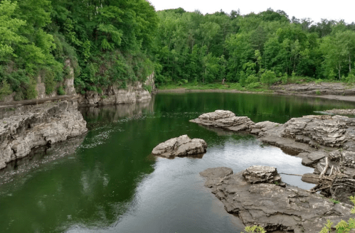 A serene green landscape featuring a calm, reflective body of water surrounded by rocky cliffs and lush trees.