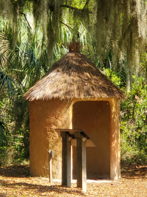 A small, round hut with a thatched roof surrounded by greenery and trees. A wooden sign stands in front.