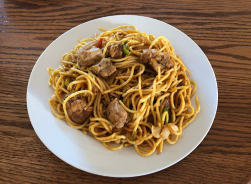 A plate of stir-fried noodles with chicken pieces and colorful vegetables on a wooden table.