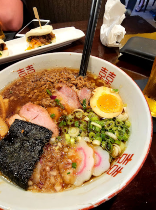 A bowl of ramen with sliced meats, an egg, seaweed, and green onions, served with a side of bao buns.
