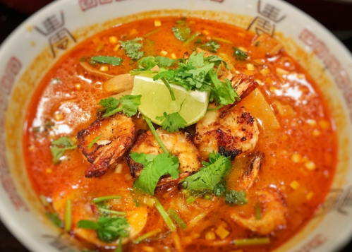 A bowl of spicy shrimp soup garnished with lime and fresh herbs, featuring vibrant colors and rich broth.