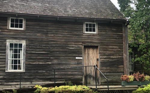 A weathered wooden house with a sloped roof, featuring multiple windows and a front door, surrounded by greenery.