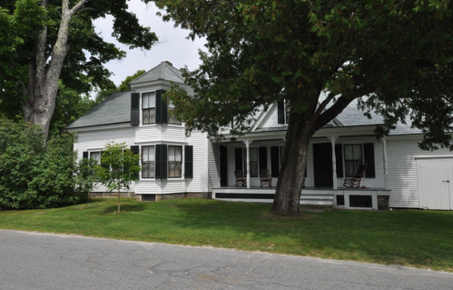 A white two-story house with black shutters, a porch, and large trees in the yard, set along a quiet street.