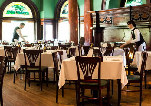 Two servers prepare a dining area with white tablecloths and elegant decor in a restaurant.