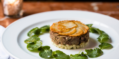 A gourmet dish featuring layered meat topped with crispy potatoes, surrounded by fresh greens on a white plate.