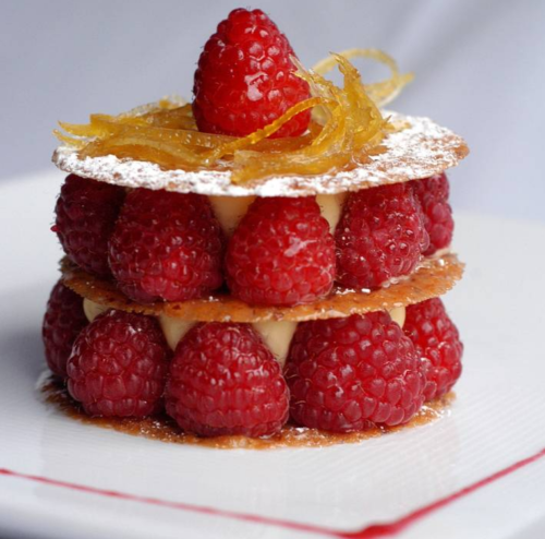 A layered dessert featuring raspberries, cream, and crispy pastry, topped with a raspberry and candied citrus peel.
