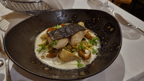 A beautifully plated dish featuring fish on a bed of vegetables in a creamy sauce, served in a dark bowl.