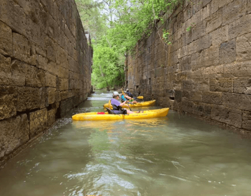 Two kayakers in yellow kayaks navigate a narrow, stone-walled waterway surrounded by lush greenery.