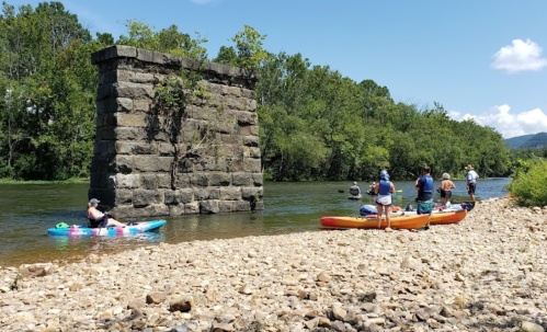 A group of kayakers near a stone pillar by a river, surrounded by lush greenery and a clear blue sky.