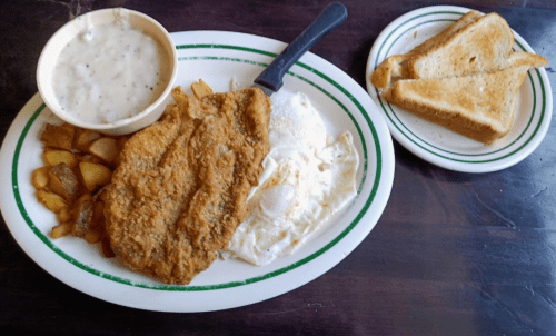 A plate with fried chicken, eggs, potatoes, and a bowl of gravy, accompanied by a side of toast.