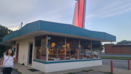 A retro-style diner with large windows, neon signage, and a woman walking by in the foreground during twilight.