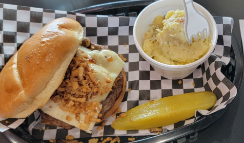 A burger with melted cheese and crispy onions, served with a side of potato salad and a pickle slice on a checkered tray.